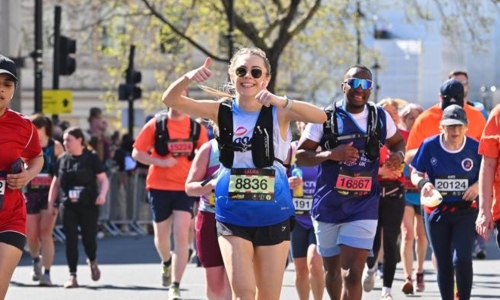 A young woman smiling with thumbs up, while running a marathon in an Age UK top