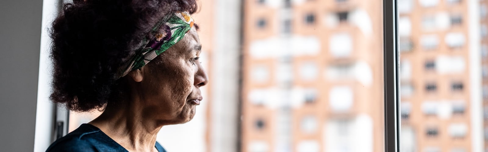 An older Black woman stares sadly out of her flat window